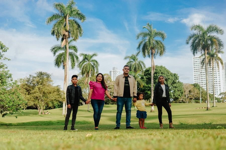 Una familia de cinco se toma de las manos en un parque con palmeras y edificios altos al fondo